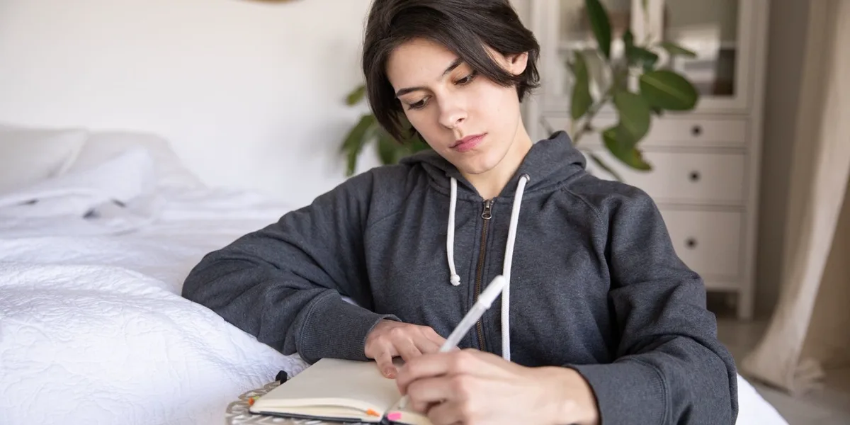A young person sitting by a bed, thoughtfully writing in a notebook